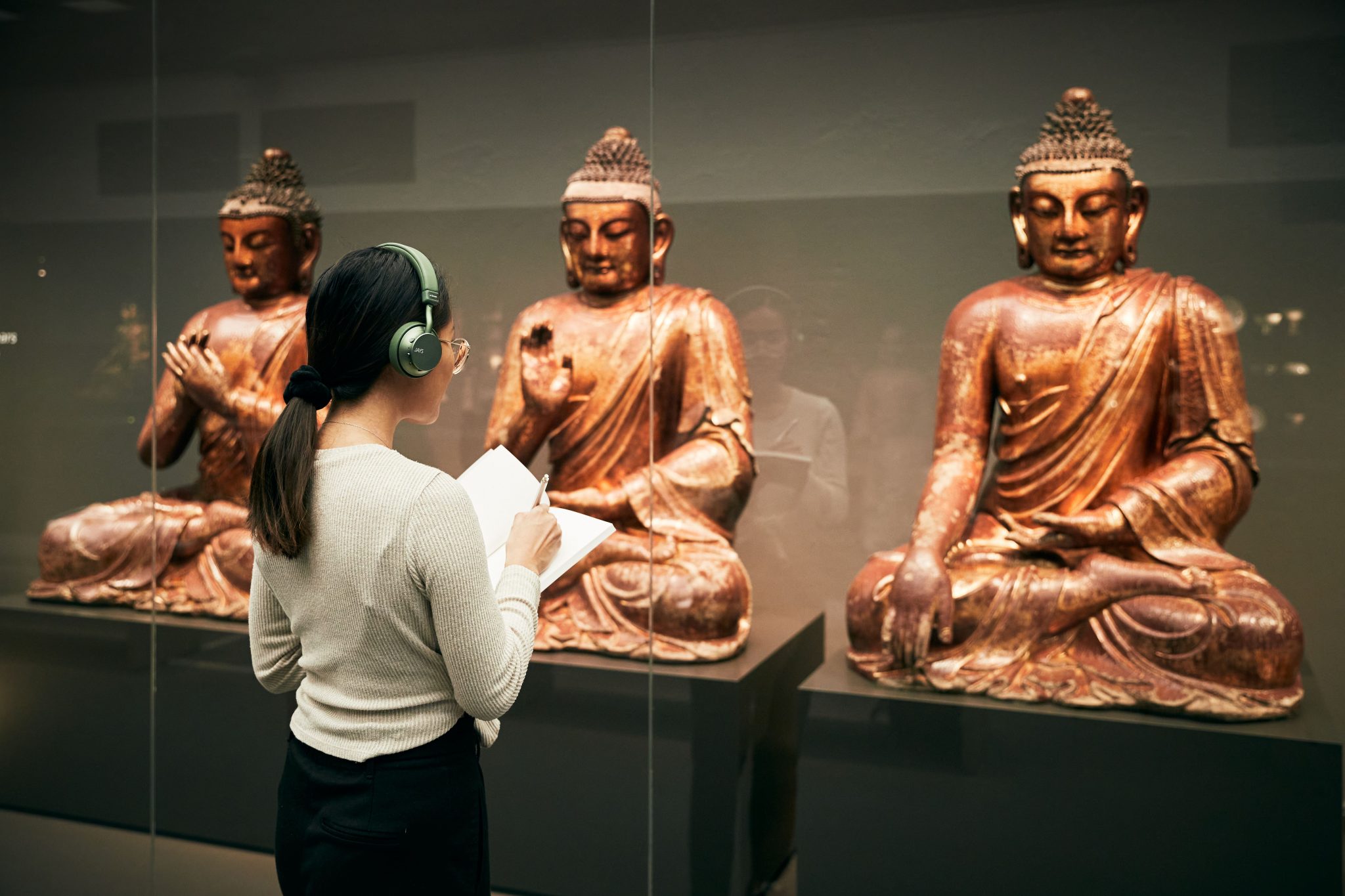 Mörkhårig kvinna med hörlurar och ett anteckningsblock står vid monter och tittar på buddhistiska statyer bakom en glasvägg