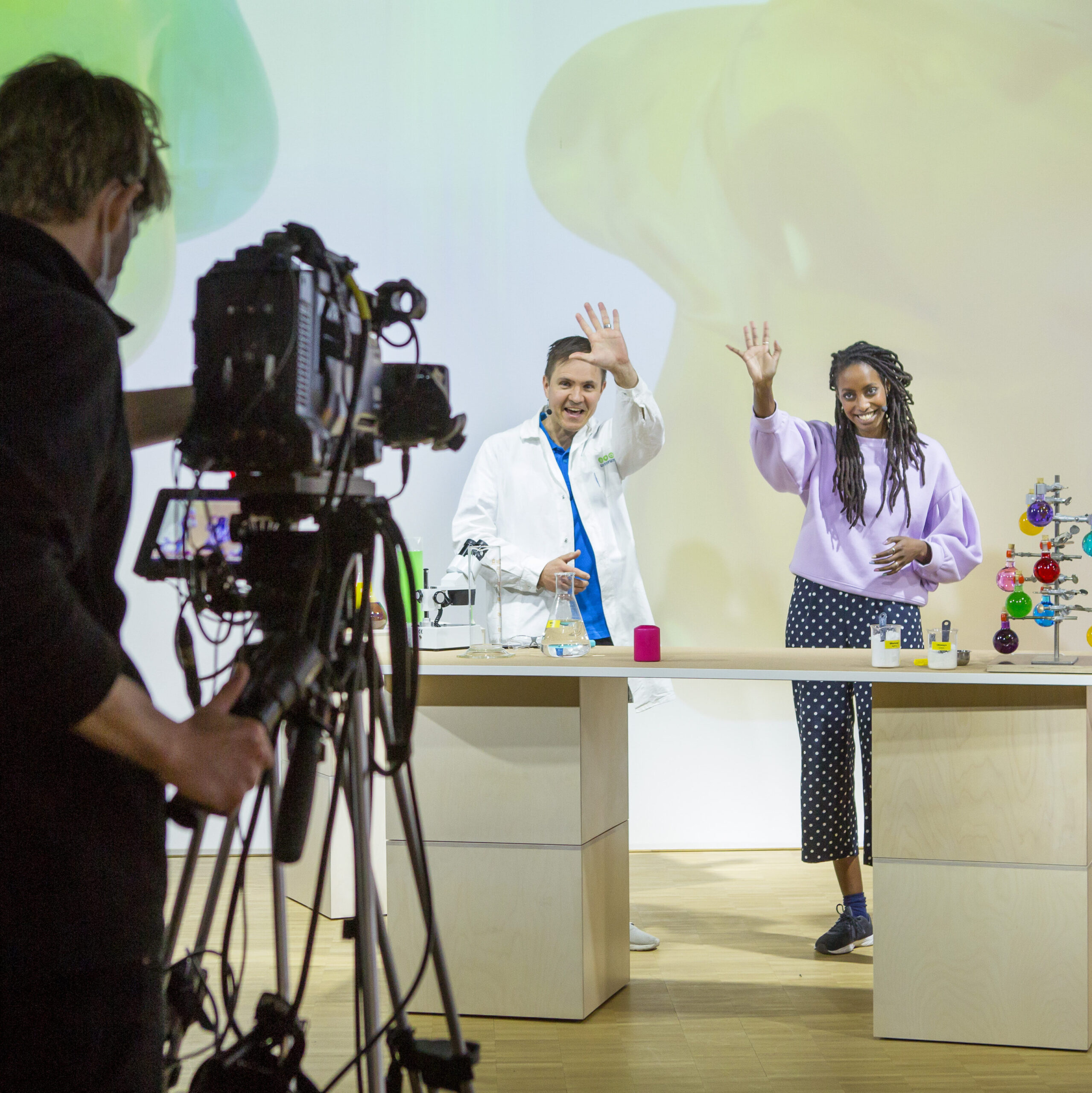 En kameraman står i en studio med en filmkamera riktad mot tre personer, iklädda vita rockar, som vinkar mot kameran, på bordet framför sig har de små flaskor med olikfärgade vätskor