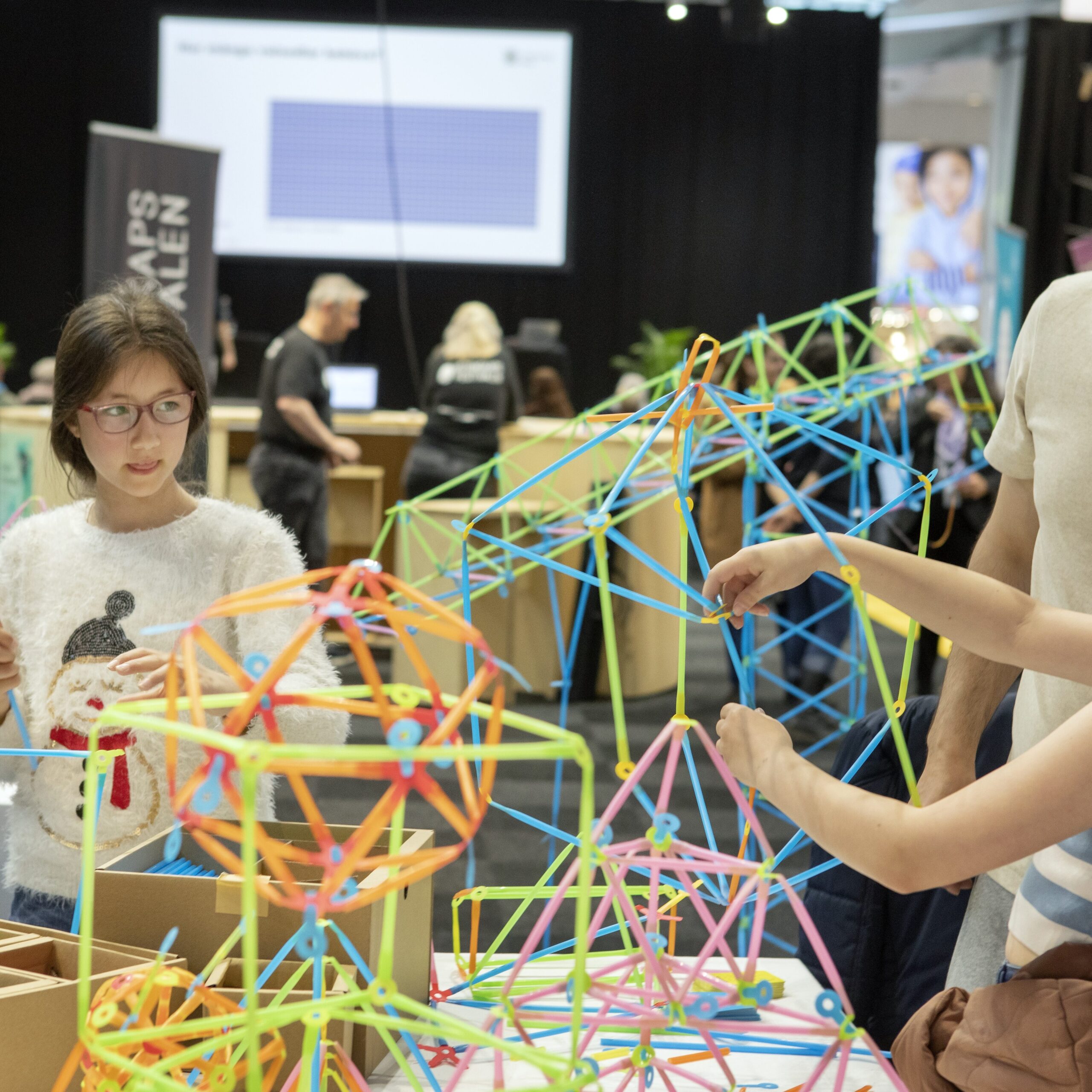 Två flickor bygger modeller av sugrör på vetnskapsfestivalen, en man hjälper till