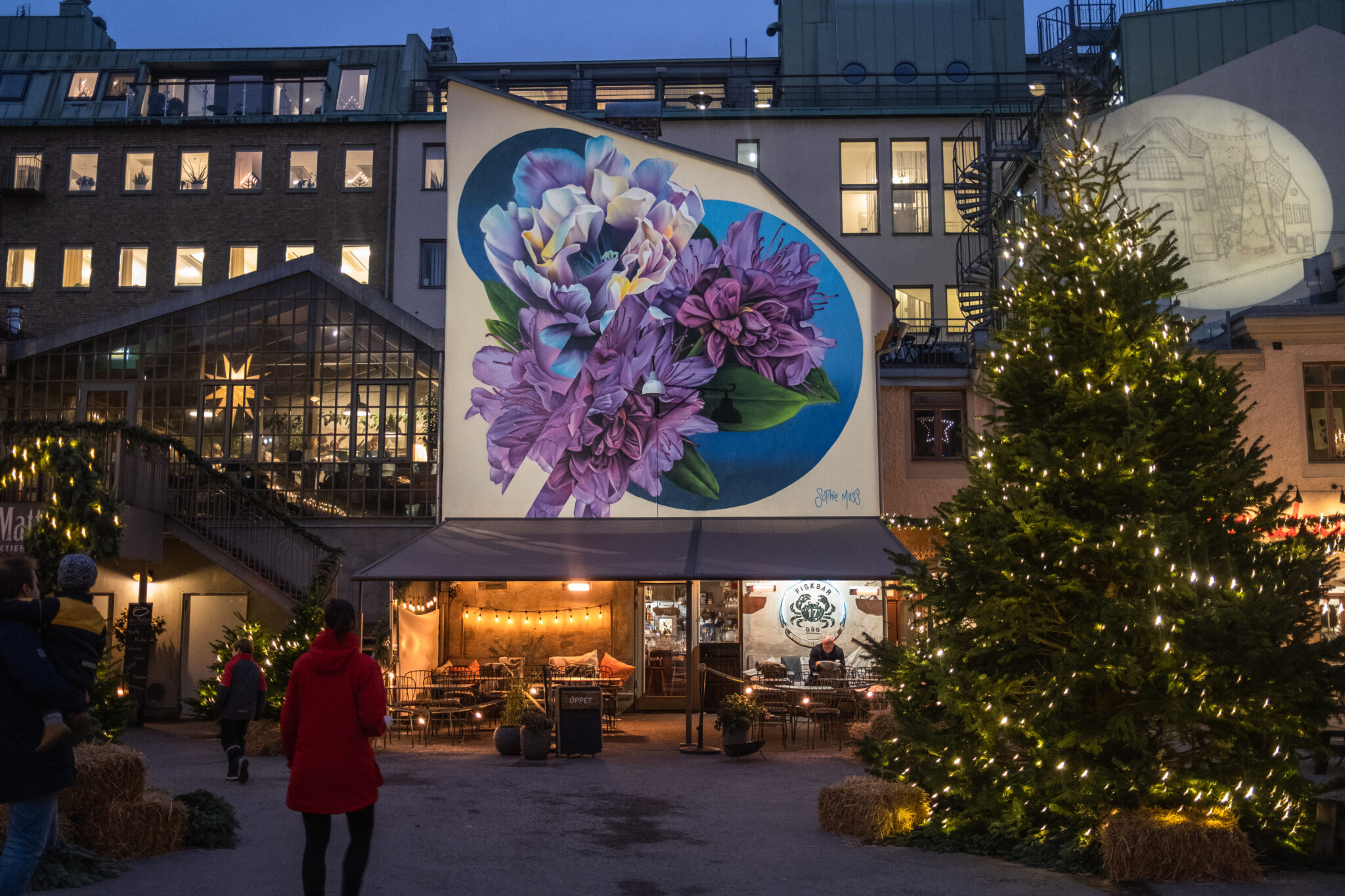 Fasadmålning med stora lila blommor på Magasinsgatan, runt om kring står julbelysta julgranar 