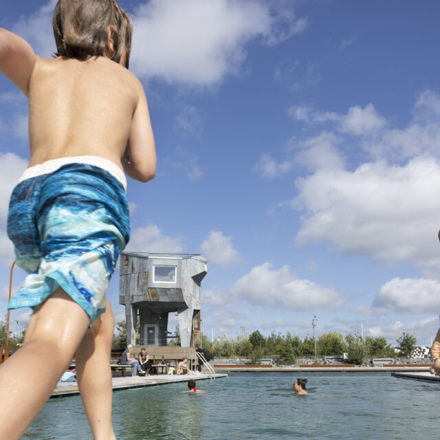 Två blonda pojkar med blå badshorts springer mot utomhuspoolen i hamnbadet vid Frihamnen Göteborg, en av dom är mitt i hoppet ner i vattnet